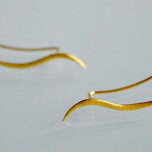 May include: A pair of gold-toned earrings with a wavy, abstract design. The earrings are made of thin metal and have a delicate, minimalist style.