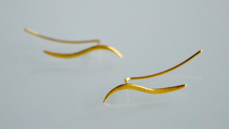 May include: A pair of gold-toned earrings with a wavy, abstract design. The earrings are made of thin metal and have a delicate, minimalist style.