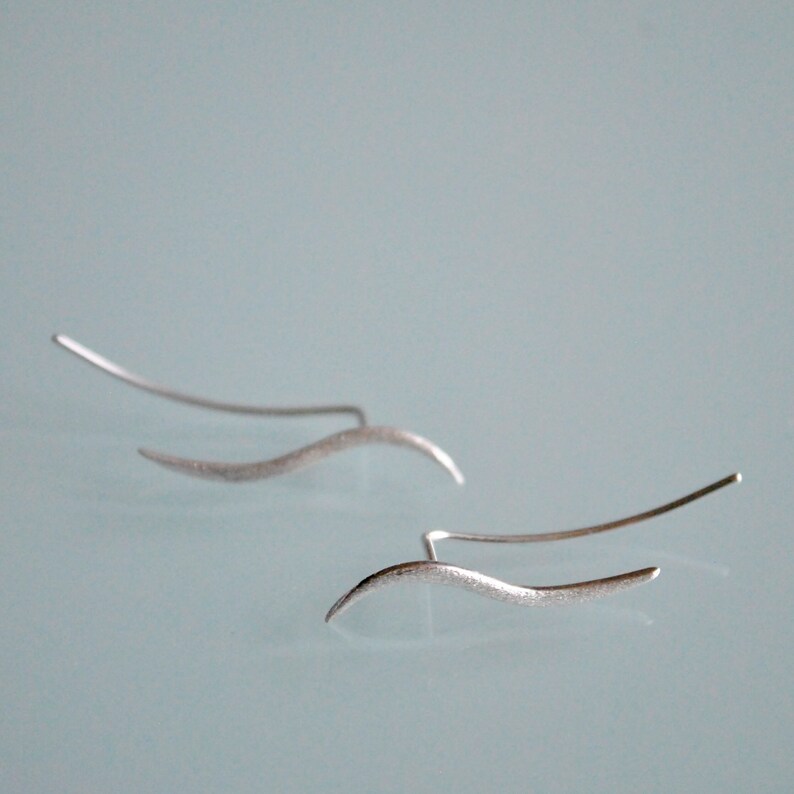 May include: A pair of silver ear cuffs with a wavy design. The cuffs are made of thin metal and have a brushed finish.