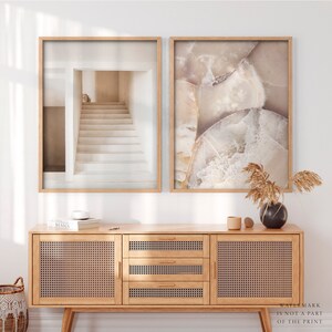 Puede incluir: Dos impresiones art&iacute;sticas enmarcadas. Una presenta una escalera minimalista en tonos neutros. La otra muestra un primer plano de una piedra con tonos beige y blanco. Las impresiones se exhiben sobre un mueble de madera clara con paneles de puerta tejidos.