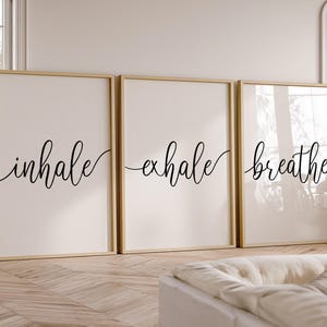 May include: Three framed prints with the words "inhale," "exhale," and "breathe" in black script. The prints have a cream background and are in gold frames. The artwork is displayed in a room with a sofa and natural light.