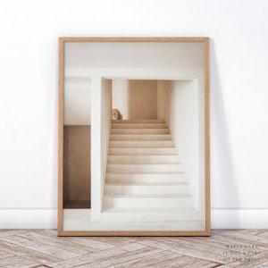 Puede incluir: Impresi&oacute;n enmarcada que presenta un interior arquitect&oacute;nico minimalista. La imagen muestra una escalera que conduce hacia arriba, ba&ntilde;ada por una luz suave y natural. La paleta de colores es neutra, con tonos beige y blanco. Un peque&ntilde;o jarr&oacute;n se encuentra en la parte superior de la escalera.