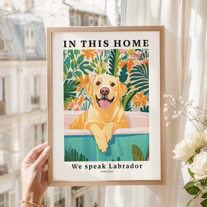 May include: A framed wall art print featuring a yellow Labrador retriever sitting in a light blue bathtub. The background displays colorful tropical leaves and flowers. The text reads IN THIS HOME at the top and We speak Labrador at the bottom.