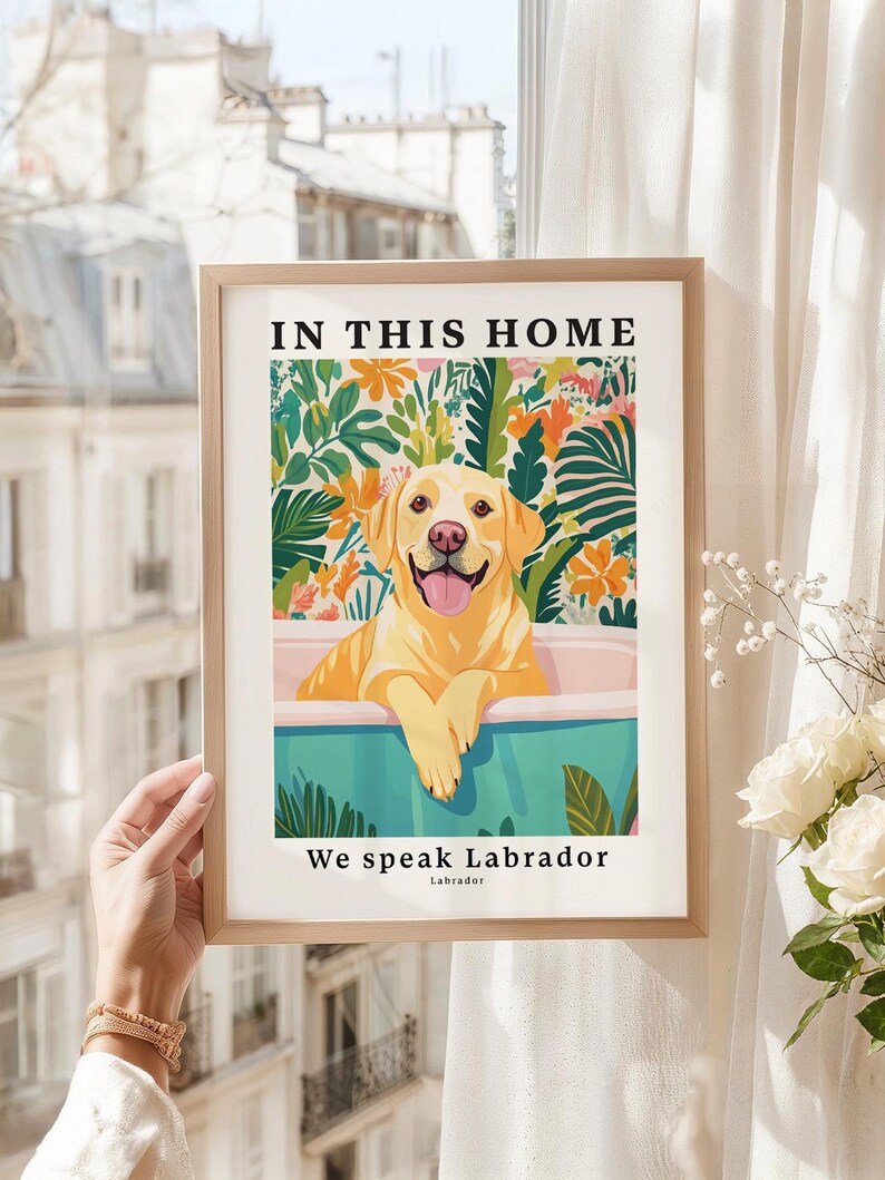 May include: A framed wall art print featuring a yellow Labrador retriever sitting in a light blue bathtub. The background displays colorful tropical leaves and flowers. The text reads IN THIS HOME at the top and We speak Labrador at the bottom.