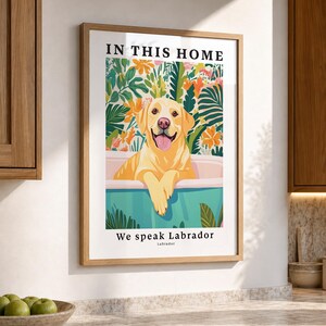 May include: A framed wall art print featuring a yellow Labrador retriever sitting in a light blue bathtub. The background displays colorful tropical leaves and flowers. The text reads IN THIS HOME at the top and We speak Labrador at the bottom.
