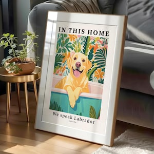 May include: A framed vertical wall art print featuring a yellow Labrador retriever sitting in a teal bathtub. The text reads IN THIS HOME at the top and We speak Labrador at the bottom. The background shows colorful tropical leaves and flowers.