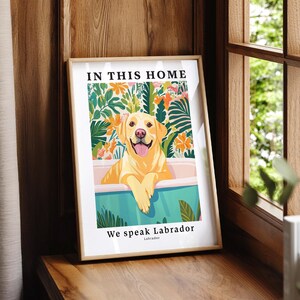 May include: A framed wall art print featuring a yellow Labrador retriever sitting in a bathtub. The background shows colorful tropical flowers and leaves. The text reads IN THIS HOME at the top and We speak Labrador at the bottom.