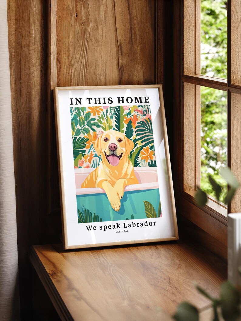 May include: A framed wall art print featuring a yellow Labrador retriever sitting in a bathtub. The background shows colorful tropical flowers and leaves. The text reads IN THIS HOME at the top and We speak Labrador at the bottom.