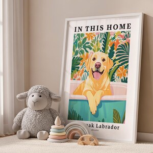 May include: A framed vertical poster featuring a yellow Labrador retriever in a bathtub surrounded by colorful tropical foliage. The text reads IN THIS HOME and below the tub, it says ak Labrador. Beside the frame is a gray plush sheep toy.