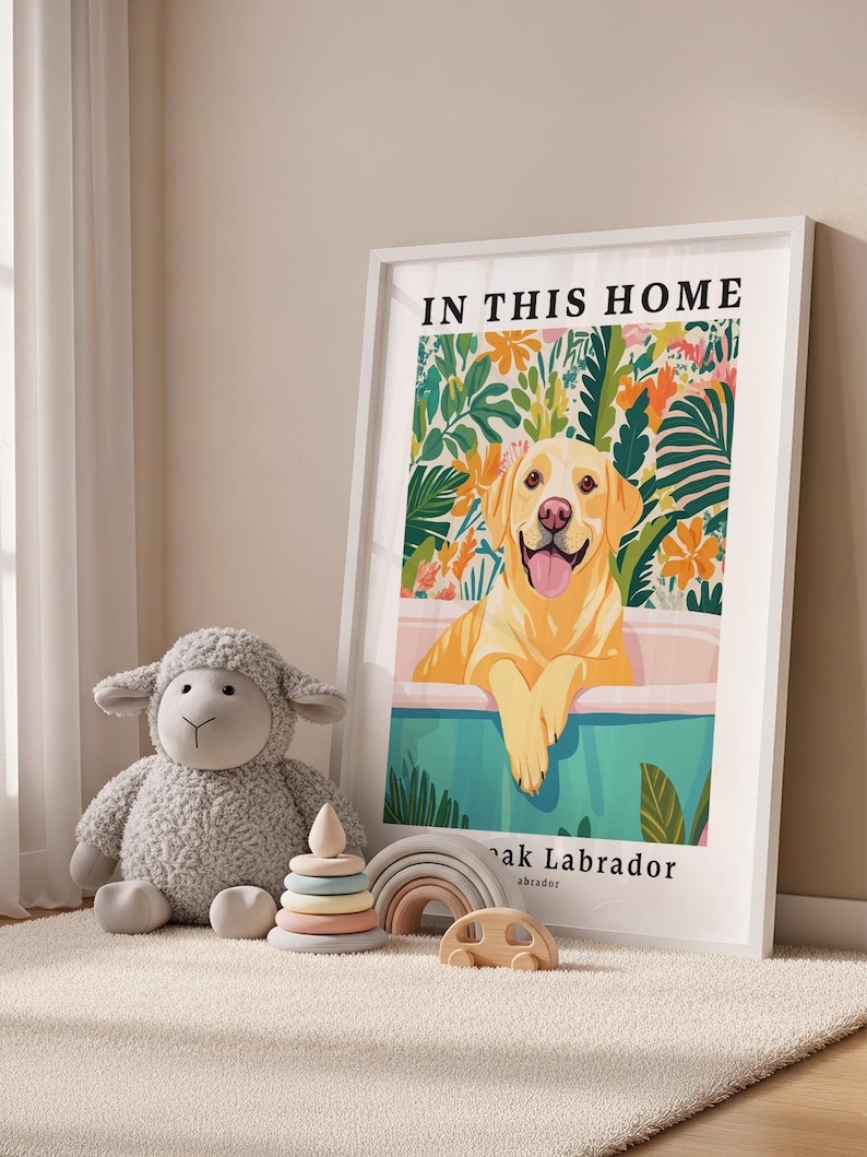 May include: A framed vertical poster featuring a yellow Labrador retriever in a bathtub surrounded by colorful tropical foliage. The text reads IN THIS HOME and below the tub, it says ak Labrador. Beside the frame is a gray plush sheep toy.