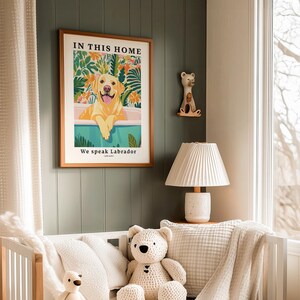 May include: A framed wall art print featuring a yellow Labrador retriever in a bathtub surrounded by colorful tropical foliage. The text reads IN THIS HOME at the top and We speak Labrador at the bottom. The print is displayed in a wooden frame.
