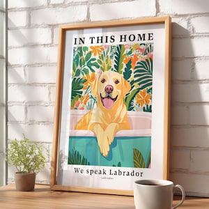 May include: A framed wall art print featuring a yellow Labrador retriever sitting in a bathtub. The illustration includes colorful tropical foliage in the background and the text "IN THIS HOME" at the top and "We speak Labrador" at the bottom.