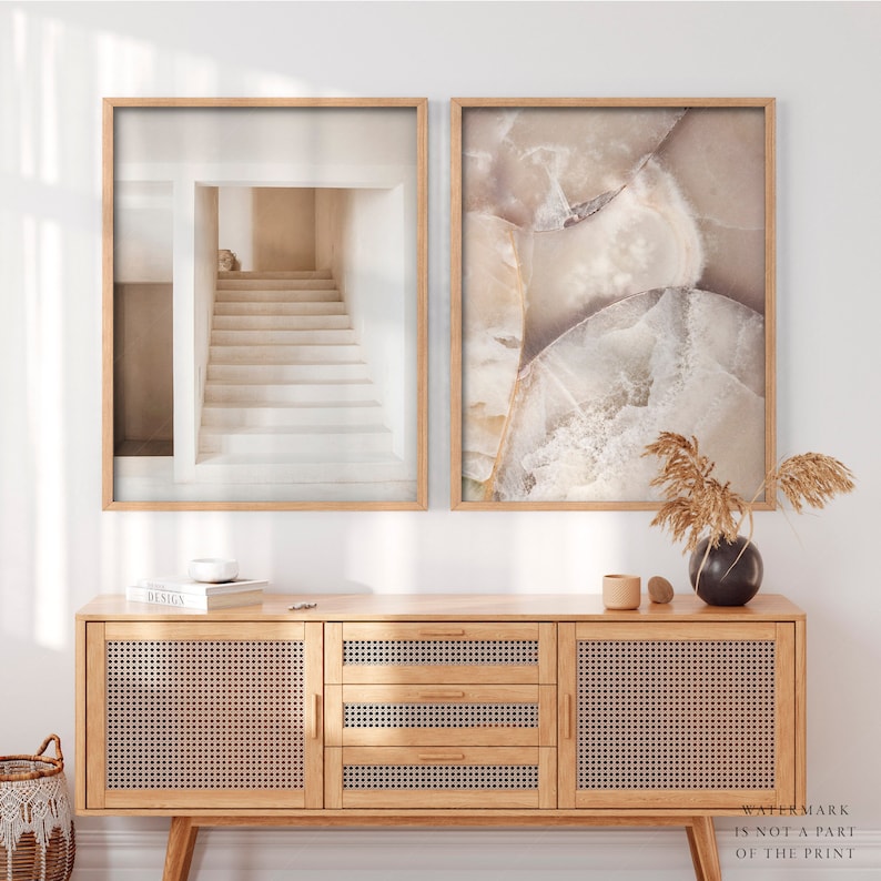 Puede incluir: Dos impresiones art&iacute;sticas enmarcadas. Una presenta una escalera minimalista en tonos neutros. La otra muestra un primer plano de una piedra con tonos beige y blanco. Las impresiones se exhiben sobre un mueble de madera clara con paneles de puerta tejidos.