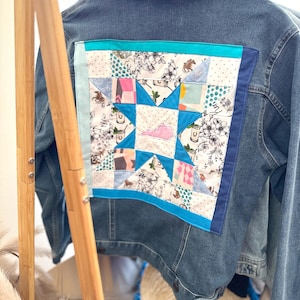 May include: A blue denim jacket with a patchwork quilt design on the back. The quilt is made of various fabrics in shades of pink, blue, white, and green. The center of the quilt features a large star with a pink outline.