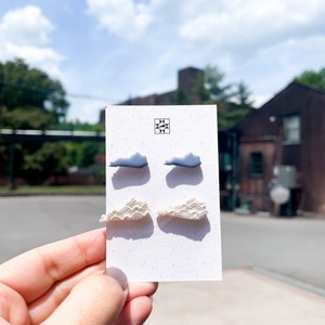 May include: A pair of earrings on a white card. The earrings are shaped like clouds, with two pairs in different colors. One pair is light blue, and the other is off-white. The card has a black and white geometric design in the upper left corner.