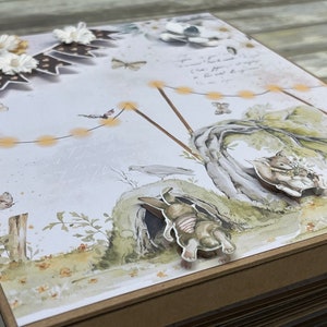 Könnte beinhalten: Ein handgemachtes Scrapbook mit einem verspielten Waldthema. Der Umschlag zeigt einen Papierhintergrund mit einer Waldszene, darunter einen Baum, einen Vogel, ein Kaninchen und eine Lichterkette. Der Umschlag ist mit Papierblumen, Schmetterlingen und einem Banner verziert.