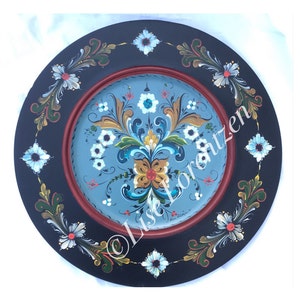 May include: A round wooden plate painted with a blue, green, yellow, and red floral design. The plate has a black border with a red inner ring.