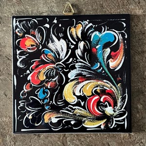 May include: Square black decorative tile with a colorful floral design. The artwork features red, yellow, blue, and white accents. A gold-colored metal triangle is attached to the top for hanging.