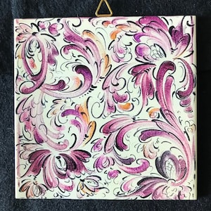 May include: A square ceramic tile with a white background and a purple and orange floral pattern. The tile has a decorative border.