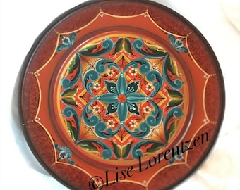 May include: A round, orange plate with a hand-painted design in shades of blue, green, red, and yellow. The design features a central flower with intricate floral patterns surrounding it. The plate has a dark brown rim and the artist's signature, "Lise Lorentzen", is visible in the lower right corner.
