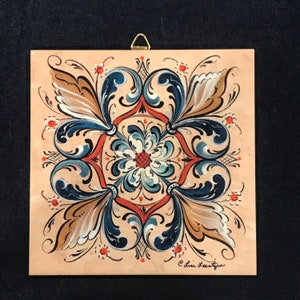 May include: Square, hand-painted decorative tile featuring a symmetrical floral design. The artwork showcases a central flower with blue and white petals, surrounded by stylized leaves in blue, brown, and white, set against a peach background. A gold hanger is attached.