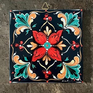 May include: Square ceramic tile with a black background and a vibrant floral design. The central flower is red with a teal center, surrounded by teal, orange, and white leaf-like patterns. A gold-colored metal hanger is attached.
