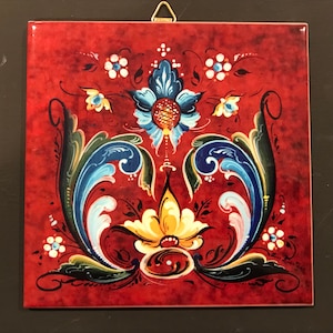 May include: Square, red decorative tile with a floral design. The design features blue, yellow, and white flowers and foliage. A gold-colored metal hanger is attached to the top.