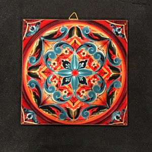 May include: Square decorative tile with a floral design. The tile features a central turquoise flower with red and white accents, surrounded by a symmetrical pattern in red, black, and turquoise. A gold-colored hook is attached to the top for hanging.
