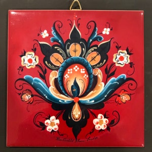 May include: A red square tile with a hand-painted floral design in black, blue, and gold. The design features a central motif of a stylized flower with a blue and gold center. The tile is signed "Lisa Crockett Flower Folk Art".