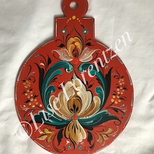 Peut inclure: Une assiette décorative en bois rouge peinte à la main avec une poignée. L'assiette présente un motif floral en turquoise, jaune et blanc. L'assiette a une bordure argentée et le texte "Lise Lorentzen".