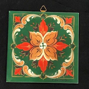 May include: Square ceramic tile with a green background and a floral design in orange, white, and brown. The design is symmetrical, with a central flower and surrounding decorative elements. A gold-colored metal hanger is attached to the top.