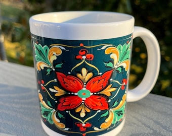 Könnte beinhalten: Weiße Keramik-Tasse mit teal-farbenem Hintergrund und einem floralen Design in Rot, Gelb und Grün. Die Tasse hat einen weißen Henkel und ein weißes Inneres. Das florale Muster ist symmetrisch und detailliert.
