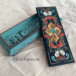 Peut inclure: Une boîte en bois peinte à la main avec un couvercle bleu sarcelle et une base noire. Le couvercle présente un motif floral dans les tons sarcelle, rouge et or. La base présente un motif floral similaire dans les tons rouge, or et blanc. La boîte est signée "Lise Lorentzen".