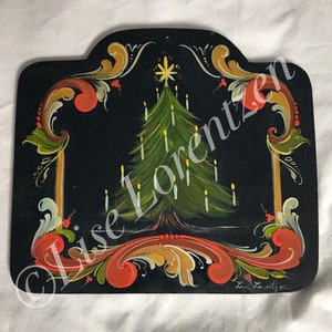以下が含まれることがあります： 黒い背景に緑色のクリスマスツリーと黄色のろうそくが描かれた、手描きの木製看板。ツリーは赤、金、緑の花柄で囲まれています。看板には「Lise Lorentzen」の署名があります。