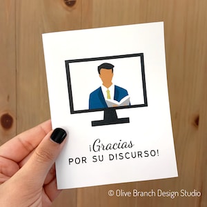 May include: A white card with a black computer monitor on it. The monitor shows a person in a blue suit and yellow tie giving a presentation. The text on the card says "&iexcl;Gracias por su discurso!" and "&copy; Olive Branch Design Studio".