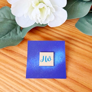 Pode incluir: Um cartão quadrado azul com um design salpicado azul claro e branco. Um quadrado menor com as letras "JW" em estilo manuscrito está centrado no cartão. Flores brancas e folhas verdes estão no fundo.