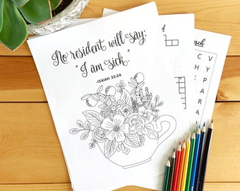 May include: White coloring pages with floral designs and the text "No resident will say: 'I am sick.' -ISAIAH 33:24". A set of colored pencils in various colors are arranged on a wooden surface.