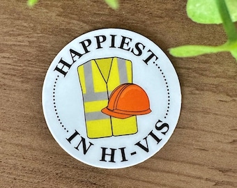 Puede incluir: Una pegatina redonda blanca con texto negro que dice "Happiest in Hi-Vis". La pegatina presenta un chaleco de seguridad amarillo con tiras reflectantes y un casco naranja.