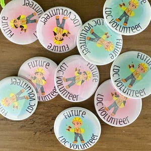 May include: A collection of round pin-back buttons featuring a cartoon character wearing a yellow hard hat and safety vest, holding a drill. The text "FUTURE LDC VOLUNTEER" is printed in a circular arc around the character on a watercolor background.