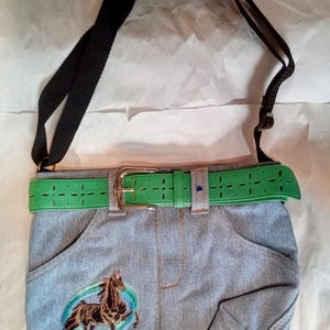 May include: A denim crossbody bag with a green belt and a horse embroidered on the front. The bag has a black adjustable strap.
