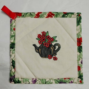 May include: A white quilted pot holder with a green and purple floral border. The center features an embroidered design of a black teapot with red flowers.