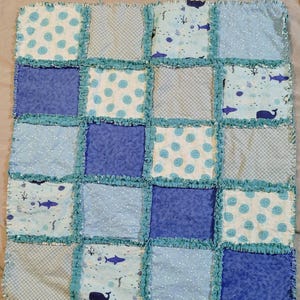 May include: A blue and white patchwork quilt with a nautical theme. The quilt is made of squares of different fabrics, some with a white background and blue polka dots, some with a blue background and white polka dots, and some with a blue background and a pattern of sea creatures. The quilt is edged with a blue ruffle.
