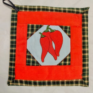 May include: Red and green quilted hot pad with a design of three red peppers on a white background. The hot pad has a black loop for hanging.