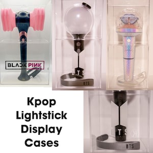 May include: Clear acrylic display cases showcasing Kpop lightsticks. The lightsticks include a pink and black hammer, a clear globe, and a holographic design. The text "BLACKPINK" and "BTS" are visible. The text "Kpop Lightstick Display Cases" is also visible.