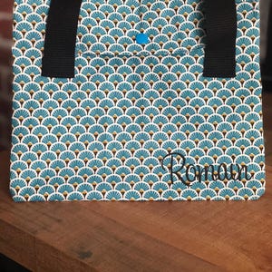 K&ouml;nnte beinhalten: Eine blaue und wei&szlig;e Einkaufstasche mit geometrischem Muster und dem Namen "Romana" auf der Vorderseite gestickt. Die Tasche hat zwei schwarze Henkel.