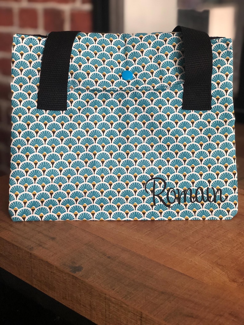 K&ouml;nnte beinhalten: Eine blaue und wei&szlig;e Einkaufstasche mit geometrischem Muster und dem Namen "Romana" auf der Vorderseite gestickt. Die Tasche hat zwei schwarze Henkel.