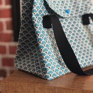 K&ouml;nnte beinhalten: Eine blaue und wei&szlig;e gemusterte Tragetasche mit schwarzen Riemen. Die Tasche hat ein geometrisches Design mit einem f&auml;cherartigen Muster.