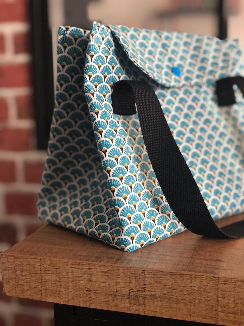 K&ouml;nnte beinhalten: Eine blaue und wei&szlig;e gemusterte Tragetasche mit schwarzen Riemen. Die Tasche hat ein geometrisches Design mit einem f&auml;cherartigen Muster.