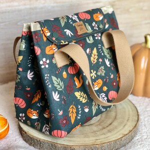 K&ouml;nnte beinhalten: Eine dunkelgr&uuml;ne Tragetasche mit einem Herbst-Motiv, das orange und rote Bl&auml;tter, K&uuml;rbisse und F&uuml;chse zeigt. Die Tasche hat einen braunen lederartigen Griff und eine kleine Tasche auf der Vorderseite mit dem Text "Yankee Fox" in goldenen Buchstaben.