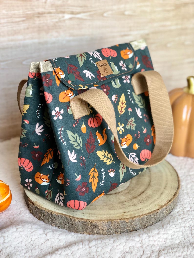 K&ouml;nnte beinhalten: Eine dunkelgr&uuml;ne Tragetasche mit einem Herbst-Motiv, das orange und rote Bl&auml;tter, K&uuml;rbisse und F&uuml;chse zeigt. Die Tasche hat einen braunen lederartigen Griff und eine kleine Tasche auf der Vorderseite mit dem Text "Yankee Fox" in goldenen Buchstaben.